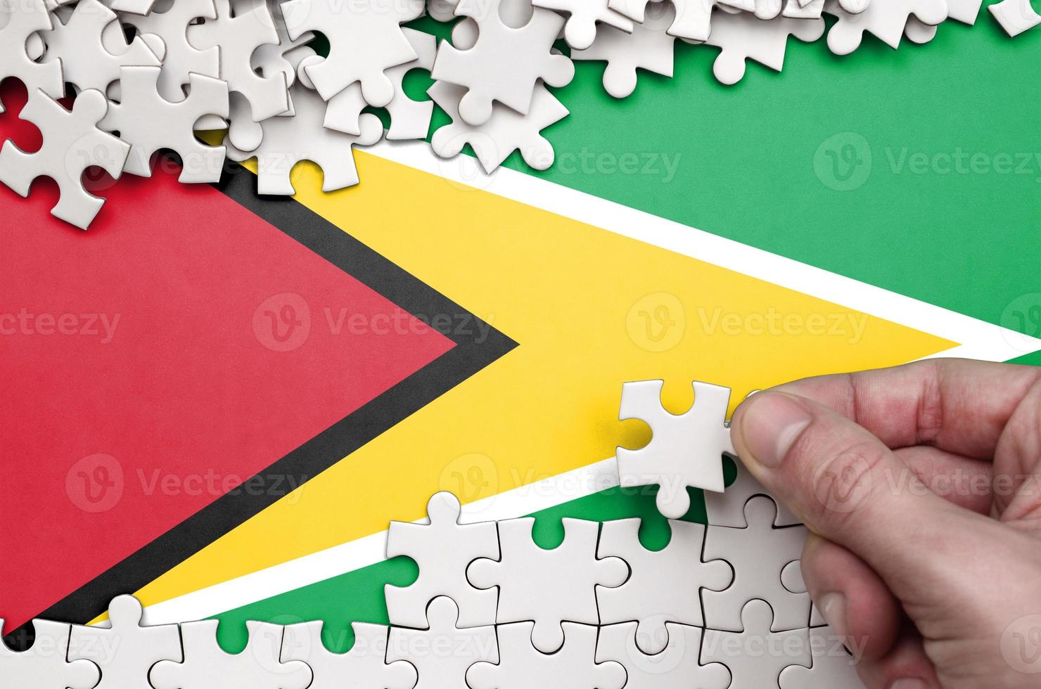 Guyana flag is depicted on a table on which the human hand folds a puzzle of white color photo