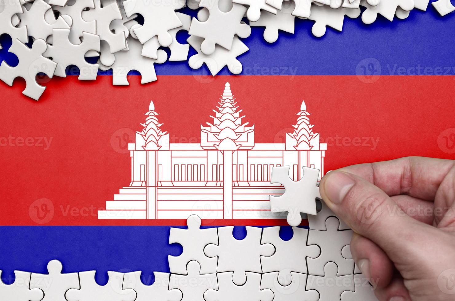 Cambodia flag is depicted on a table on which the human hand folds a puzzle of white color photo