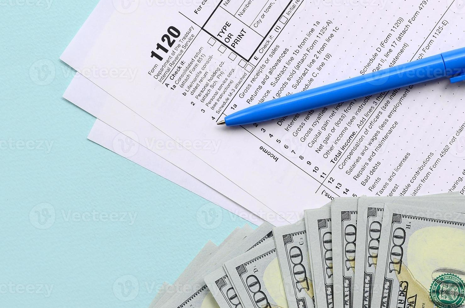 El formulario de impuestos 1120 se encuentra cerca de los billetes de cien dólares y el bolígrafo azul sobre un fondo azul claro. declaración de impuestos sobre la renta de la corporación estadounidense foto