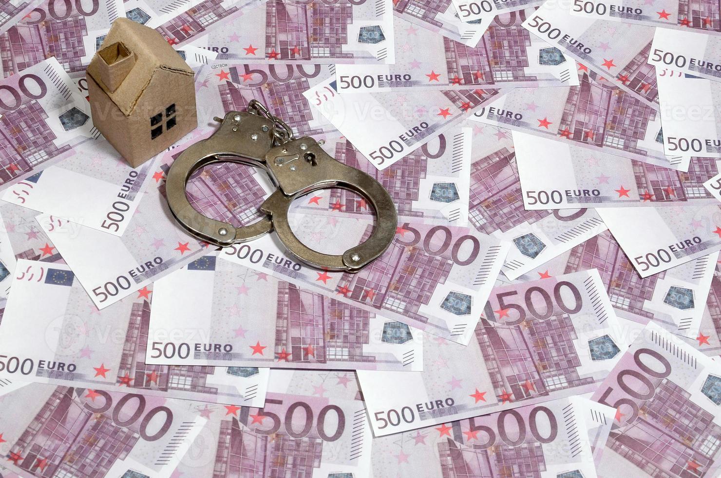Scam with real estate. Cheating when buying or renting a house. Punishment for deception. A toy house with handcuffs on a many 500 purple euro banknotes photo