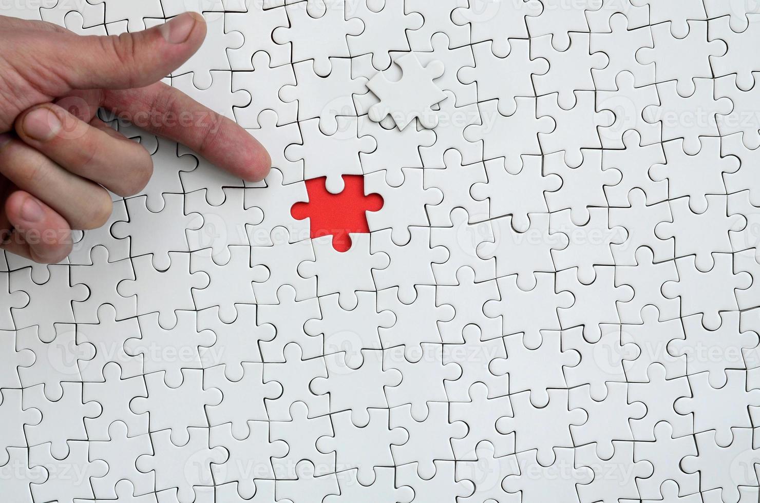 The texture of a white jigsaw puzzle in the assembled state with one missing element, forming a red space, pointed to by the finger of the male hand photo