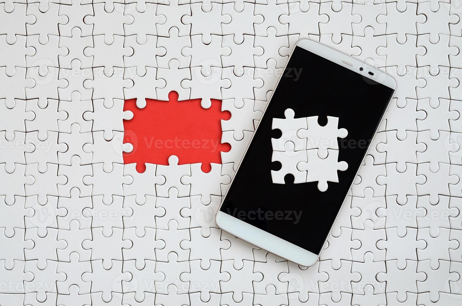 A modern big smartphone with several puzzle elements on the touch screen lies on a white jigsaw puzzle in an assembled state with missing elements photo