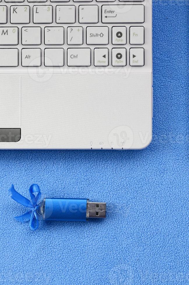 Brilliant blue usb flash memory card with a blue bow lies on a blanket of soft and furry light blue fleece fabric beside to a white laptop. Classic female gift design for a memory card photo