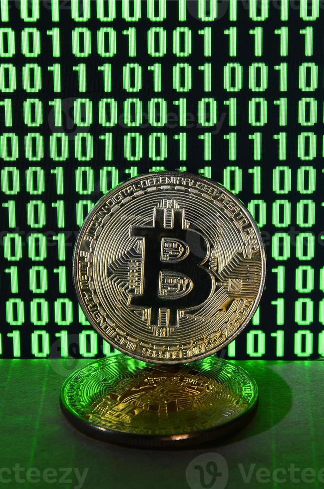 A pair of bitcoins lies on a cardboard surface on the background of a monitor depicting a binary code of bright green zeros and one units on a black background. Low key lighting photo