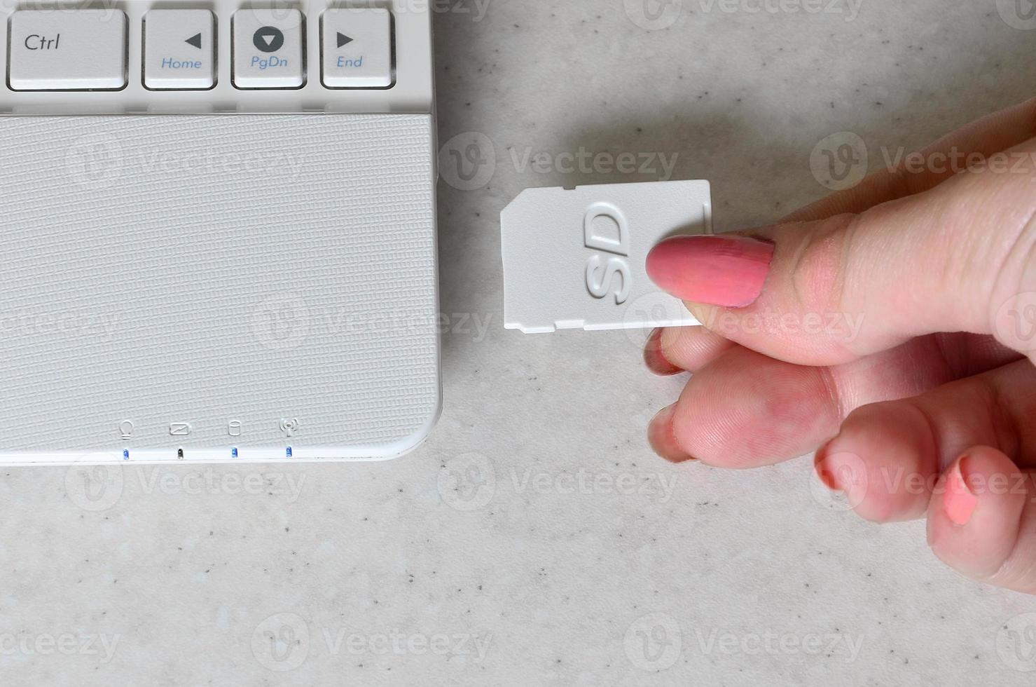 A female hand inserts a white compact SD card into the corresponding input in the side of the white netbook. Woman uses modern technologies to store memory and digital data photo