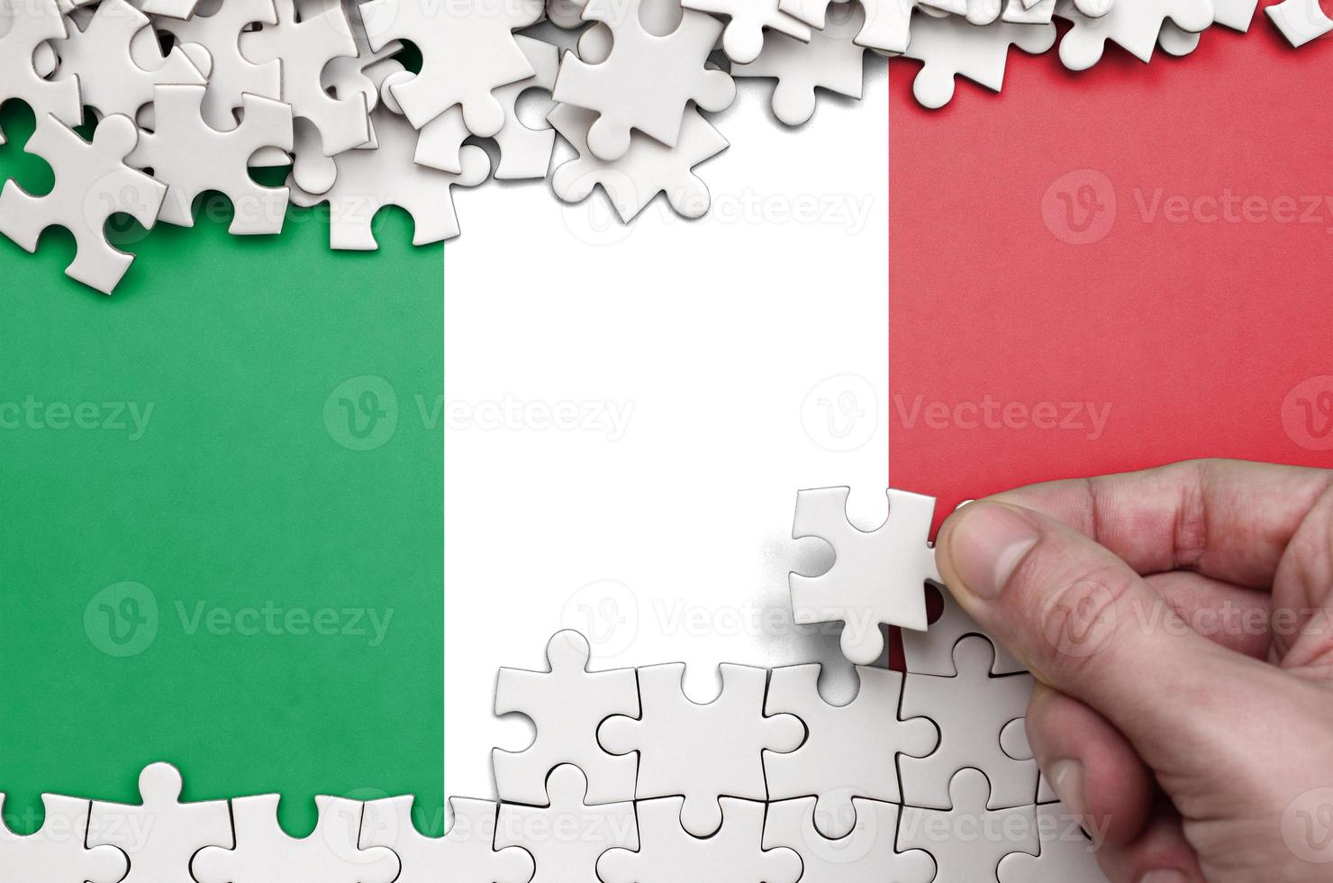 Italy flag is depicted on a table on which the human hand folds a puzzle of white color photo