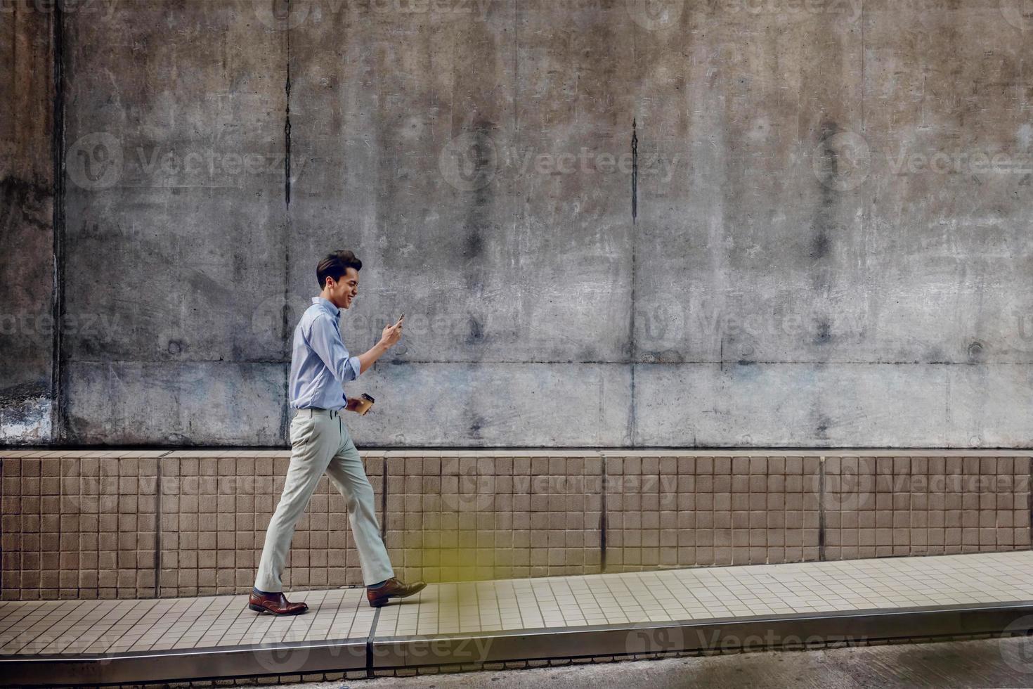 sonriente joven hombre de negocios asiático en ropa casual usando teléfono móvil mientras camina por la pared del edificio urbano. estilo de vida de la gente moderna. vista lateral. longitud total foto