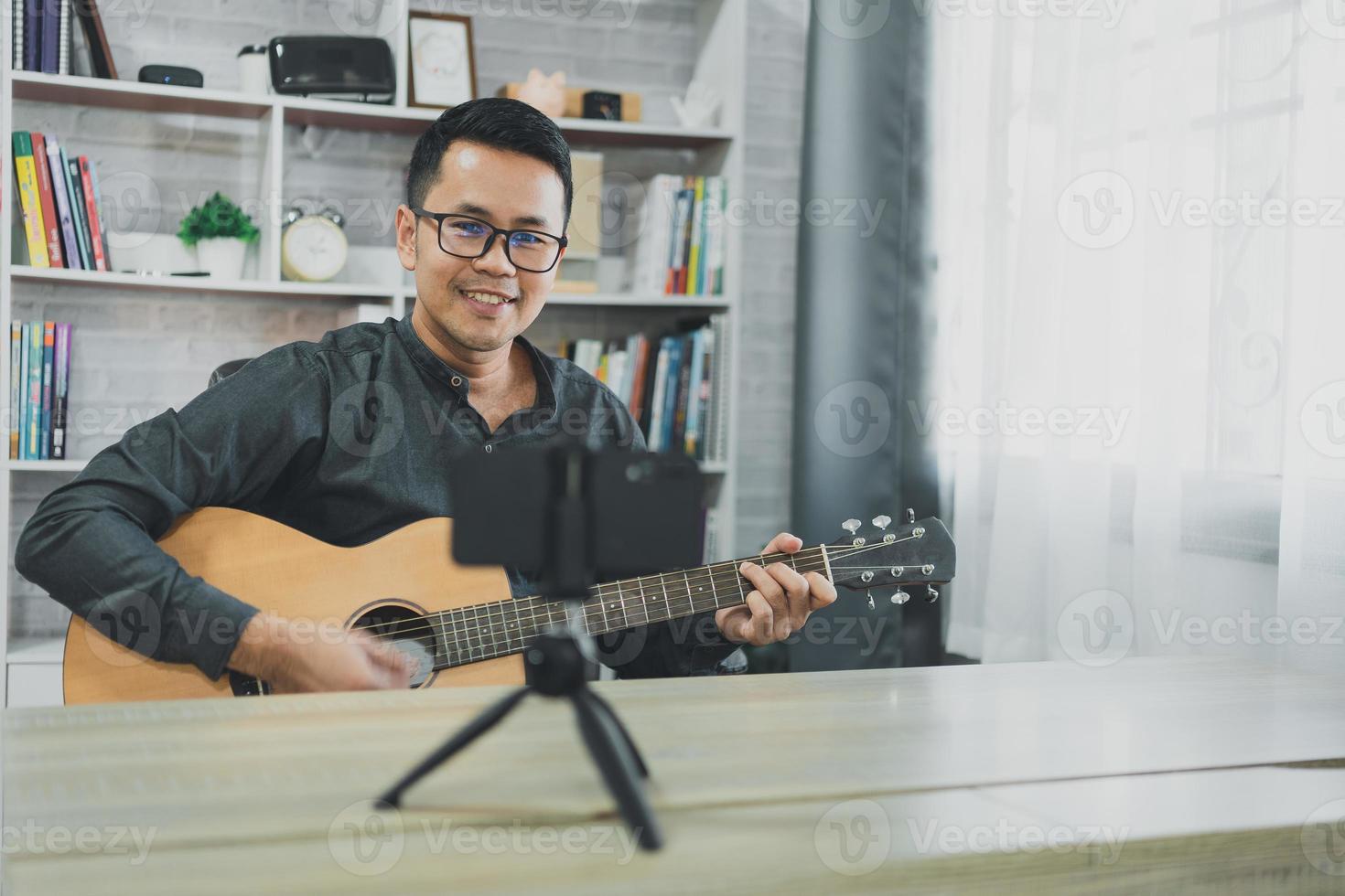 un profesor de música asiático transmite lecciones de guitarra en vivo a los estudiantes en línea usando el móvil. músico masculino asiático tocando guitarra cantando videoconferencias en vivo en casa. música de concierto en vivo desde cualquier lugar. foto