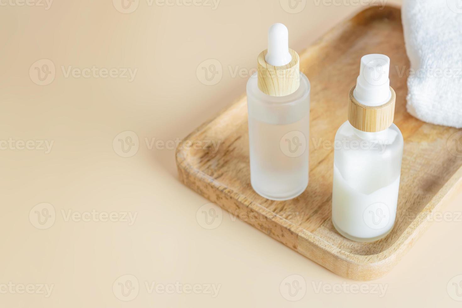 Set of skin care cosmetics - cream pump bottle and essense bottle with beauty serum or fluid on wooden plate and spa towel on beige background with copy space photo