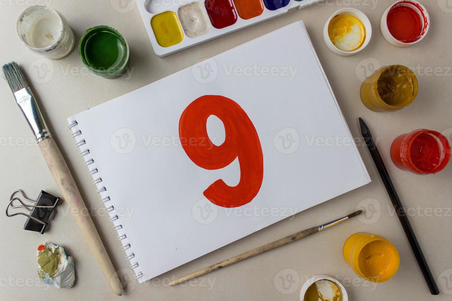 Red number nine drawing on a white album sheet. To indicate the place, price, etc. Nearby are brushes, jars of paint gouache, watercolor, etc photo