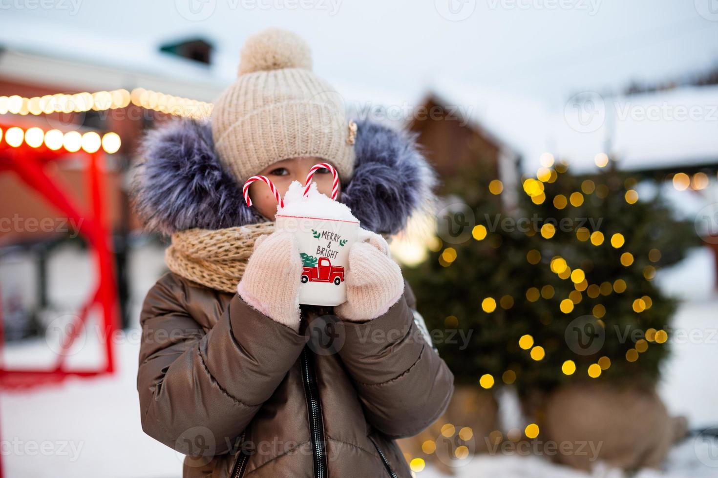 chica con taza con nieve, bastón de caramelo e inscripción alegre y brillante en sus manos al aire libre con ropa de abrigo en invierno en el mercado festivo. Guirnaldas de luces de hadas decoradas ciudad de nieve para año nuevo. Navidad foto