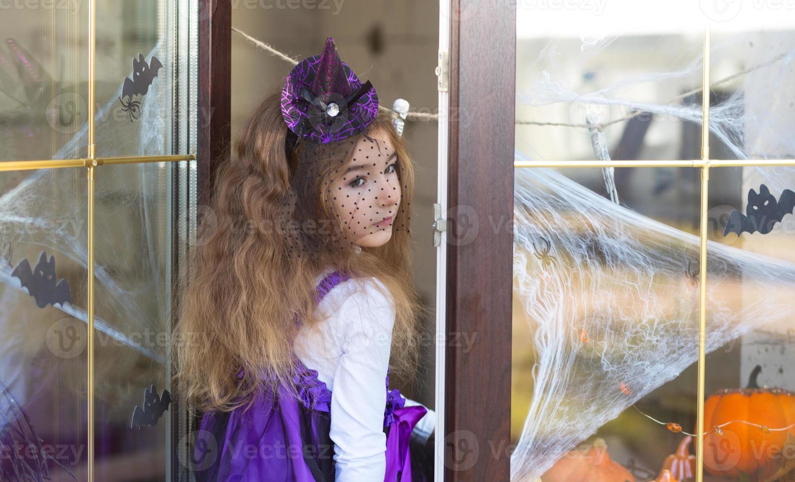 A girl in a witch costume on the windowsill by the window is playing with a spider web and a spider, decorating the house for the Halloween holiday. Terrible scenery, fear and horror, pumpkins photo