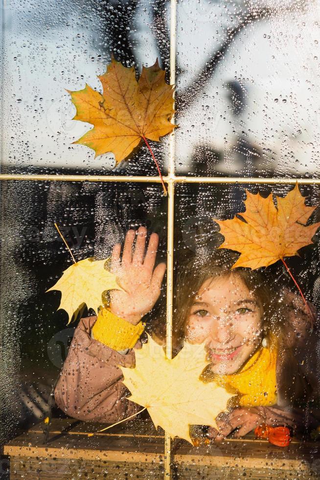 el niño mira por la ventana de la casa afuera, clima otoñal, vidrio mojado con gotas después de la lluvia, hojas de arce amarillas pegadas a la ventana. estado de ánimo otoñal, comodidad en el hogar foto