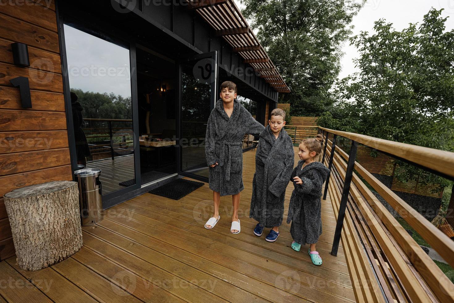 Three kids in bath robe stand in wooden terrace of modern house. photo