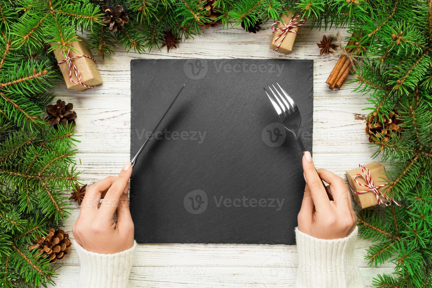 top view girl holds fork and knife in hand and is ready to eat. Empty black slate square plate on wooden christmas background. holiday dinner dish concept with new year decor photo