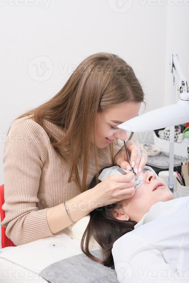young beautician woman making false lash procedure for a client. vertical shot of lash extension procedure photo