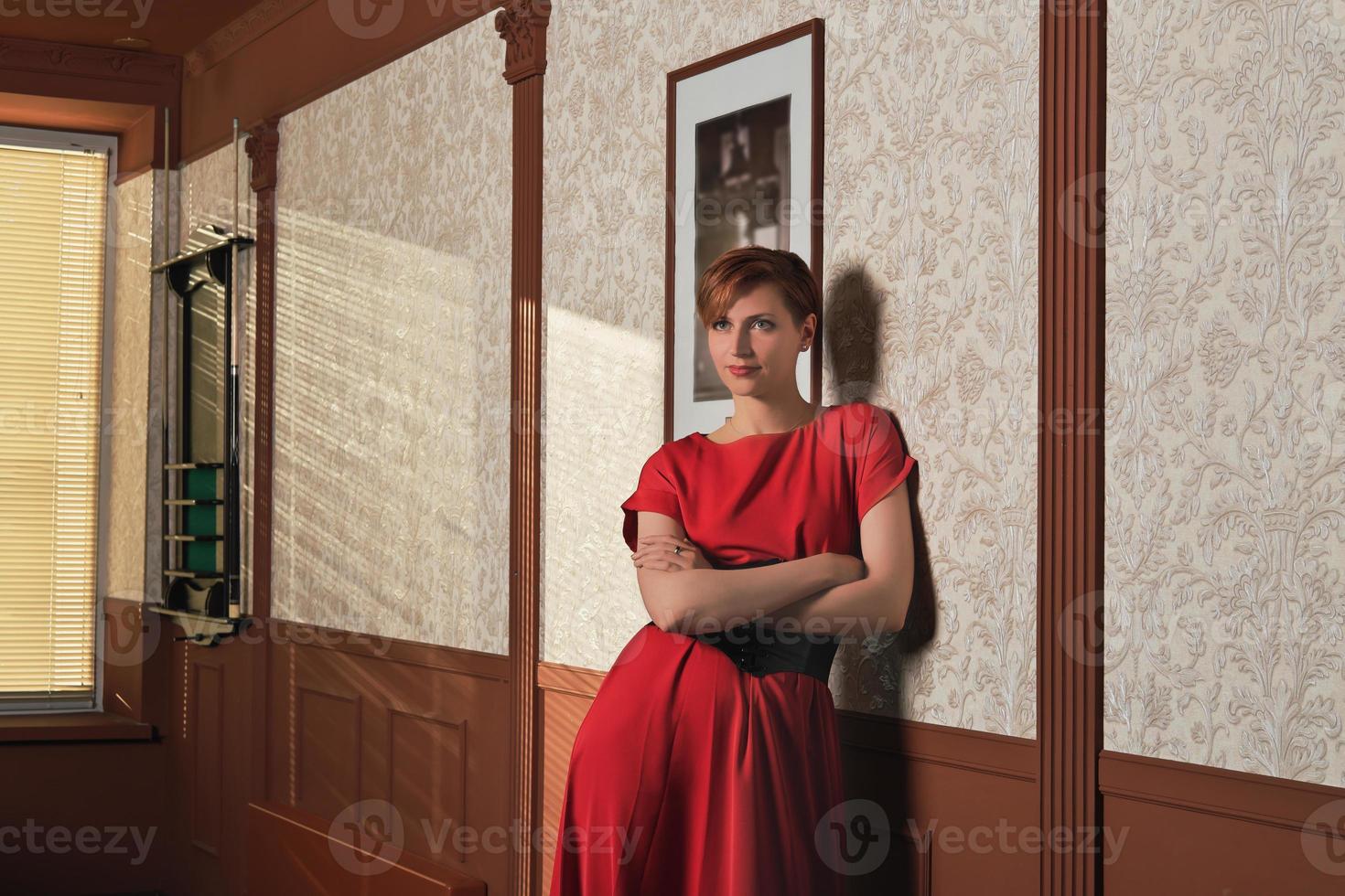 portrait of glamourous woman standing at wall in billiard pool club with hands crossed on chest. woman wearing elegant red dress looking aside photo