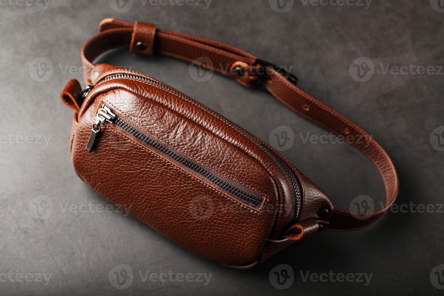 A brown belt bag made of textured brown leather on a black stone background. Elegant fanny pack brown bag with a zipper photo