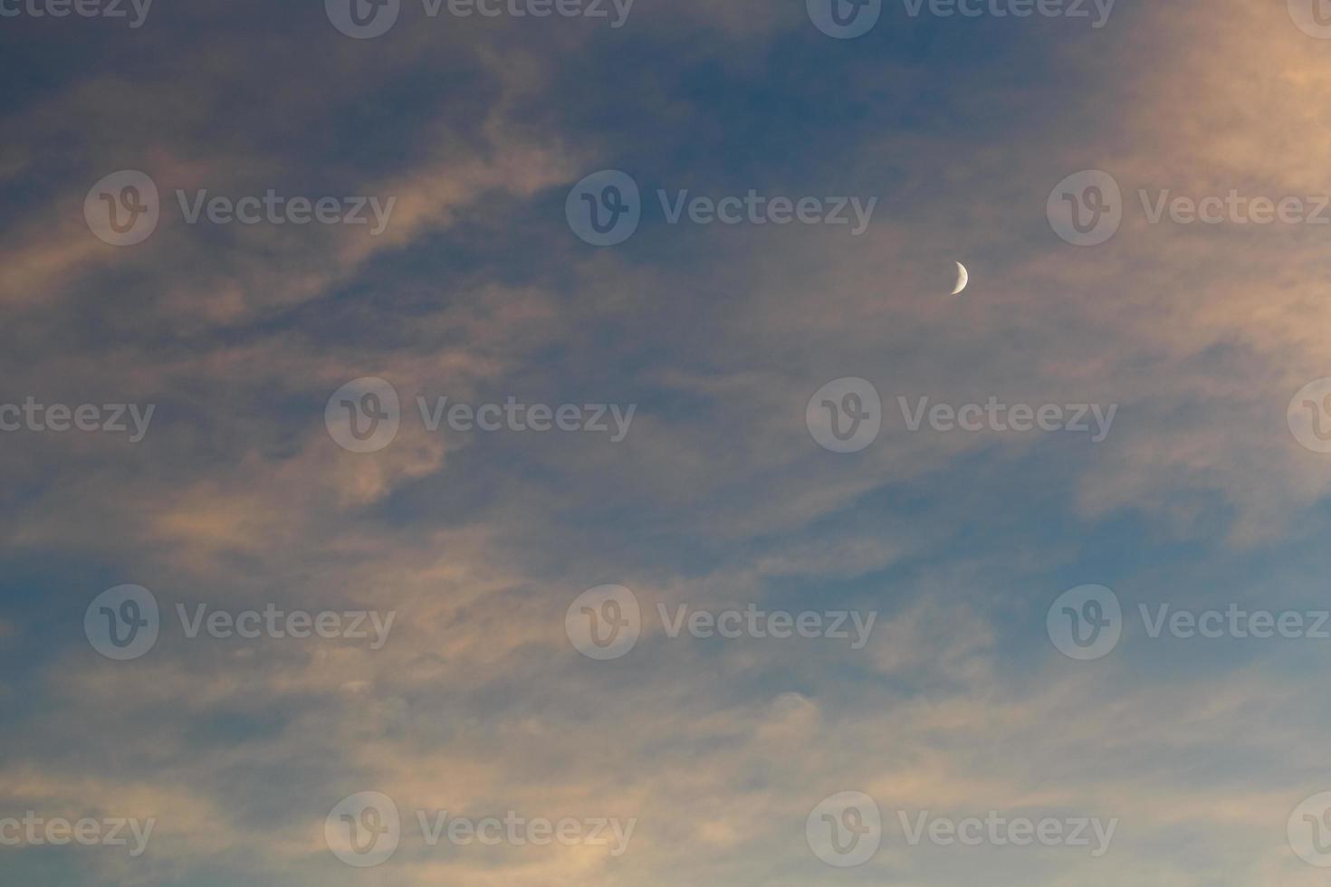 A growing moon in the blue sky among the clouds illuminated by the sun at sunset photo