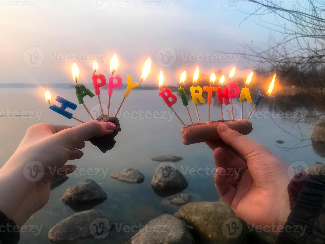 Burning happy birthday inscription made of holiday candles in the hands of a man and a woman opposite the water of the ocean lake river. Concept birthday celebration in nature, outdoors photo