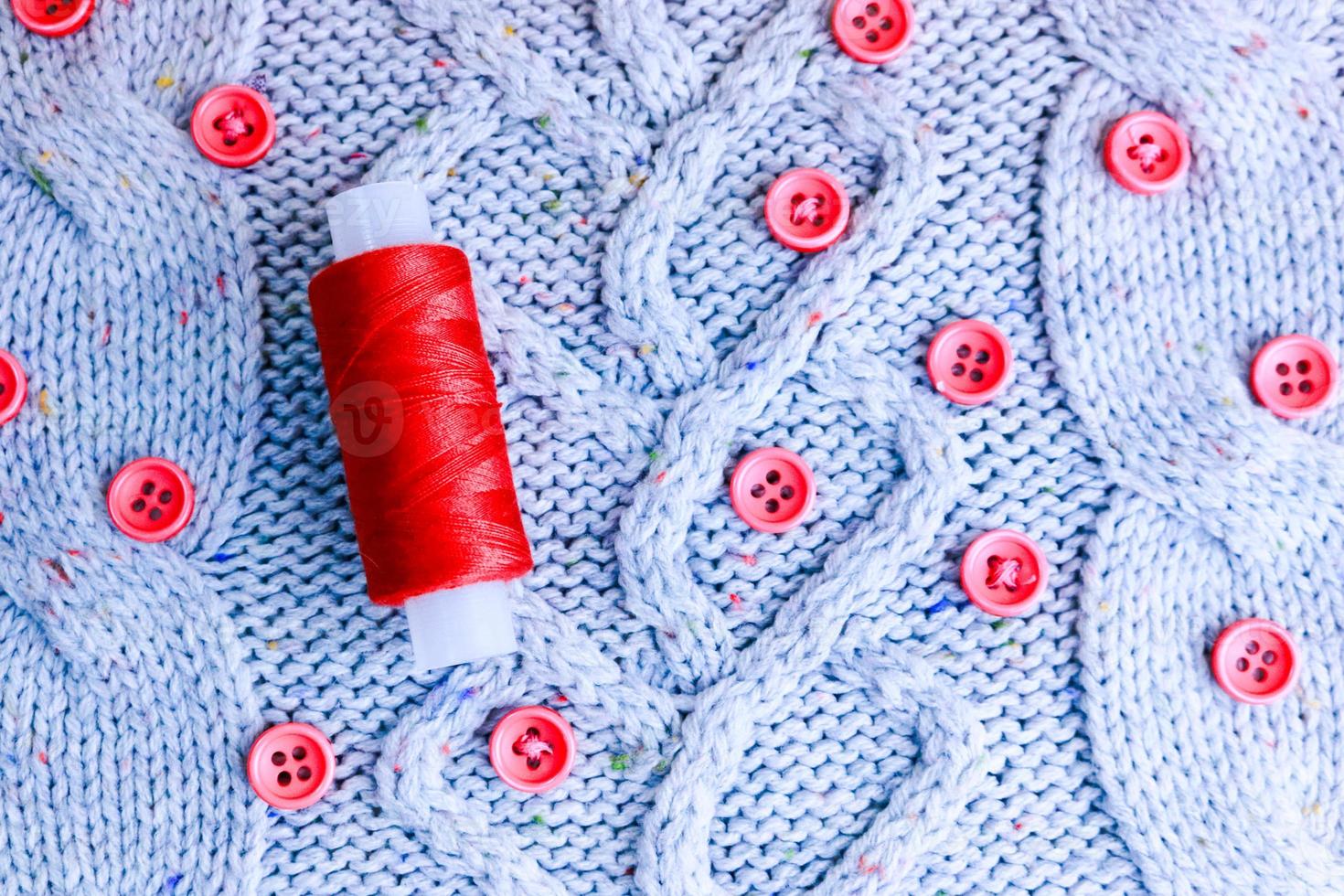 Beautiful texture of a soft warm natural sweater, fabrics with a knitted pattern of yarn and red small round buttons for sewing and a skein of red thread. The background photo