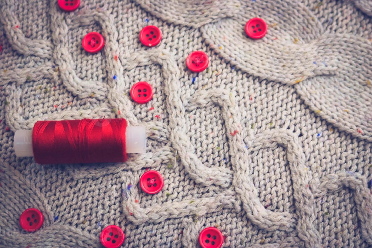 Beautiful texture of a soft warm natural sweater, fabrics with a knitted pattern of yarn and red small round buttons for sewing and a skein of red thread. The background photo