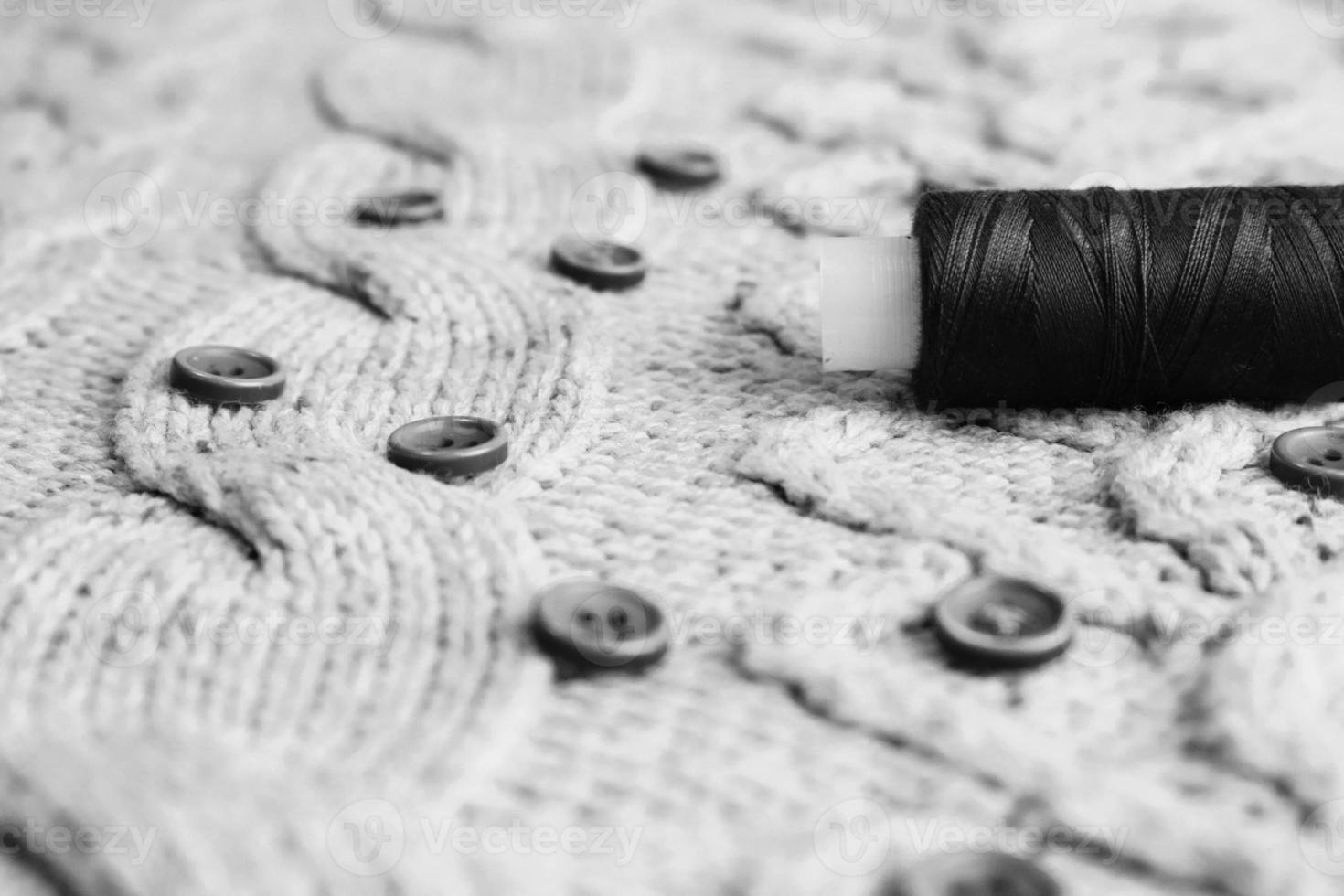 Beautiful black and white texture of a soft warm natural sweater, fabrics with a knitted pattern of threads and red small round buttons for sewing and a skein of thread. The background photo