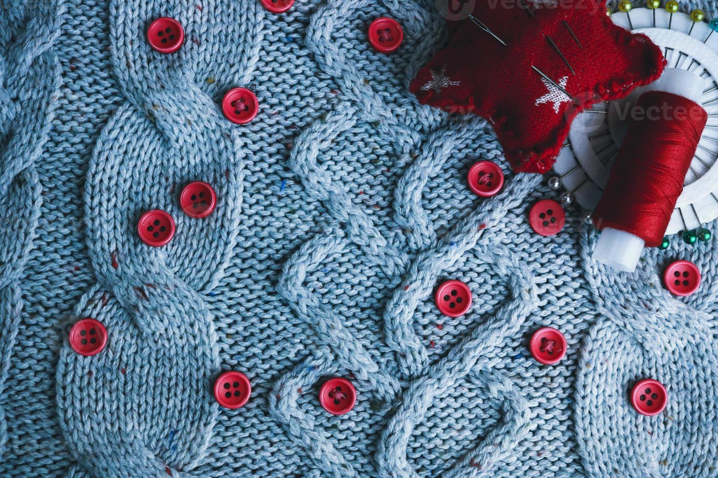 Beautiful texture of a soft warm natural sweater, fabrics with a knitted pattern and red small round buttons for sewing and a skein of thread, needle bed and needle pad. Flat lay. The background photo
