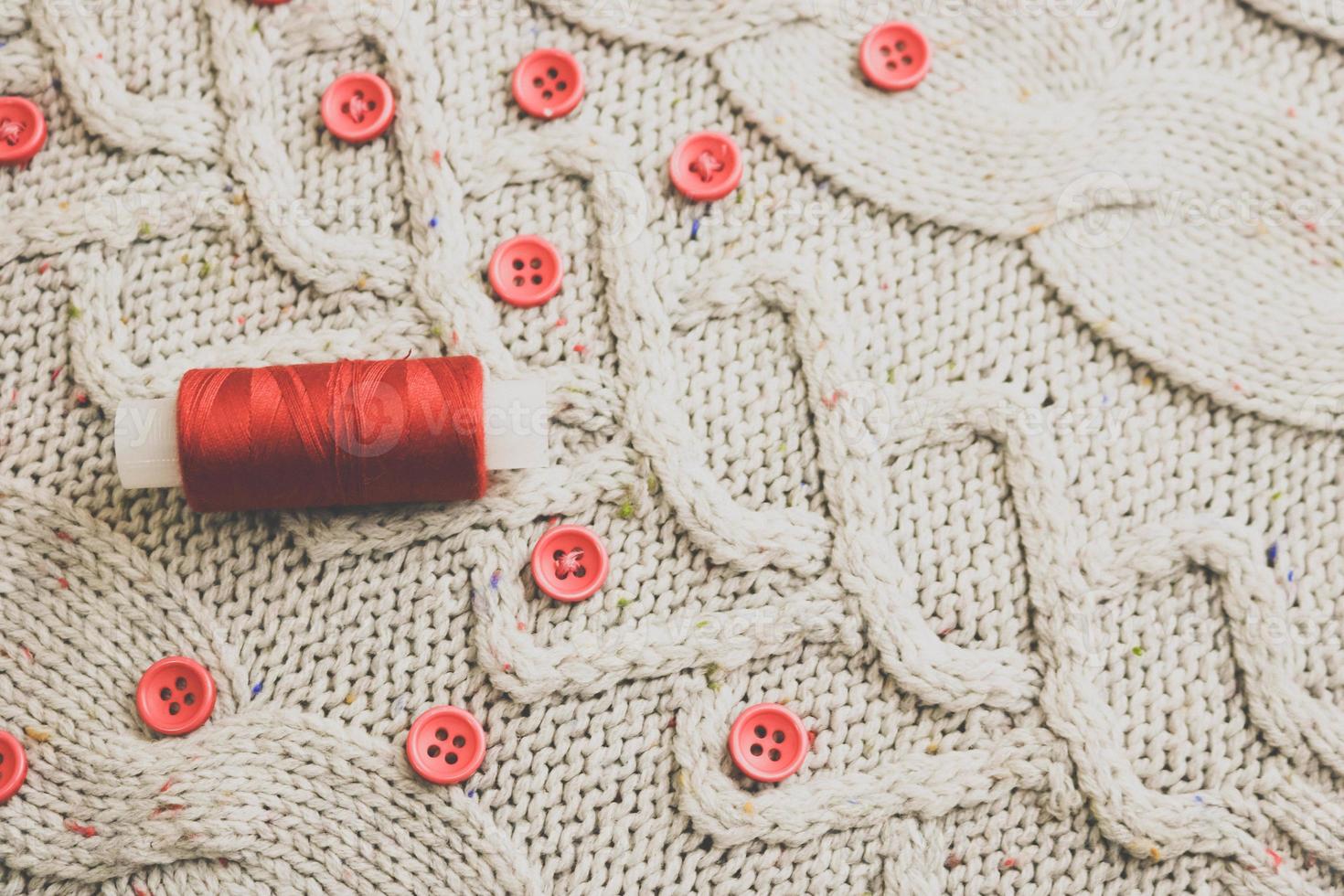 Beautiful texture of a soft warm natural sweater, fabrics with a knitted pattern of yarn and red small round buttons for sewing and a skein of red thread. The background photo