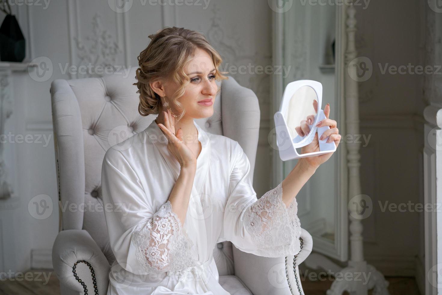 Beautiful blond woman, wearing white silk bathrobe, holding mirror and sitting photo