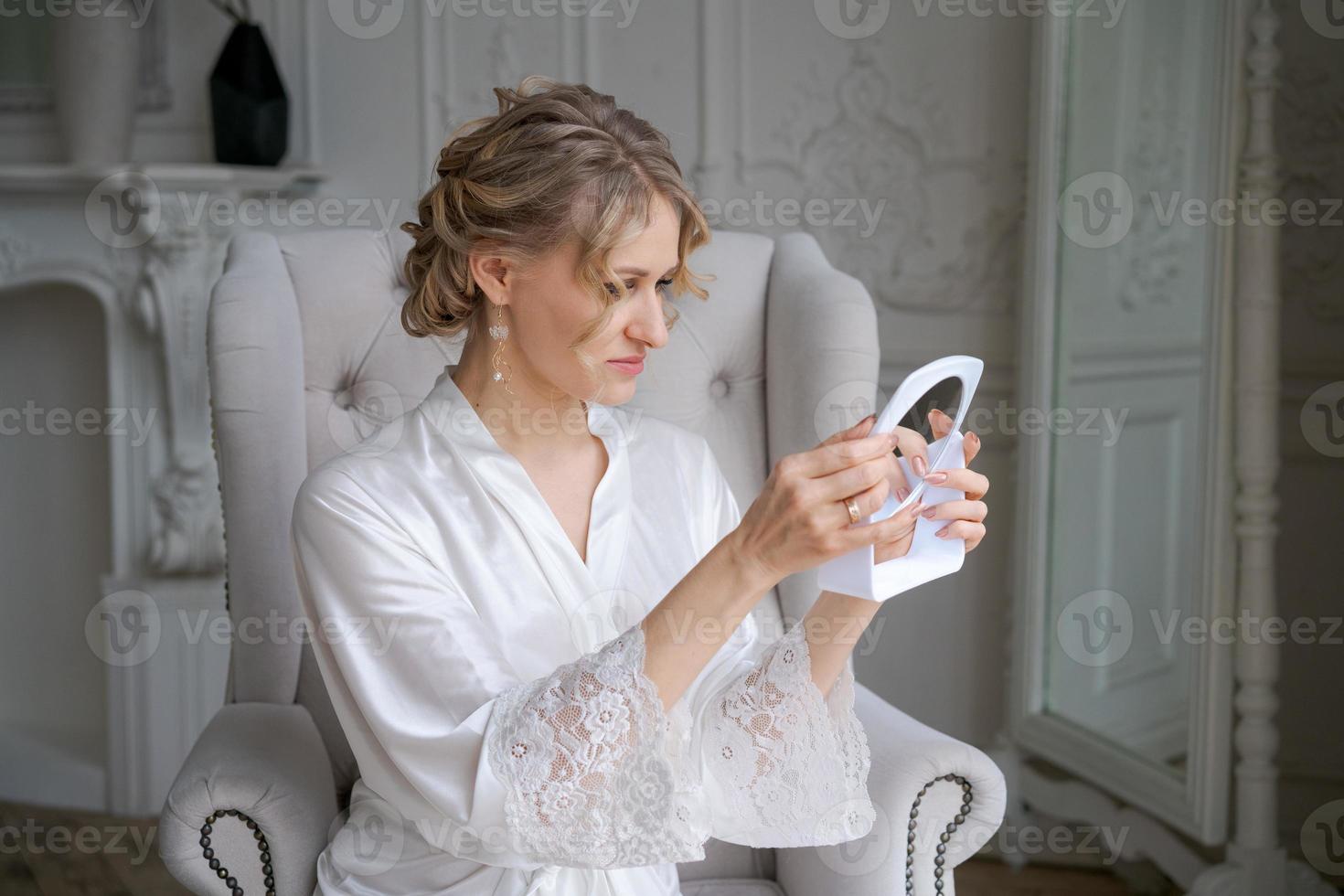Beautiful blond woman, wearing white silk bathrobe, holding mirror and sitting photo