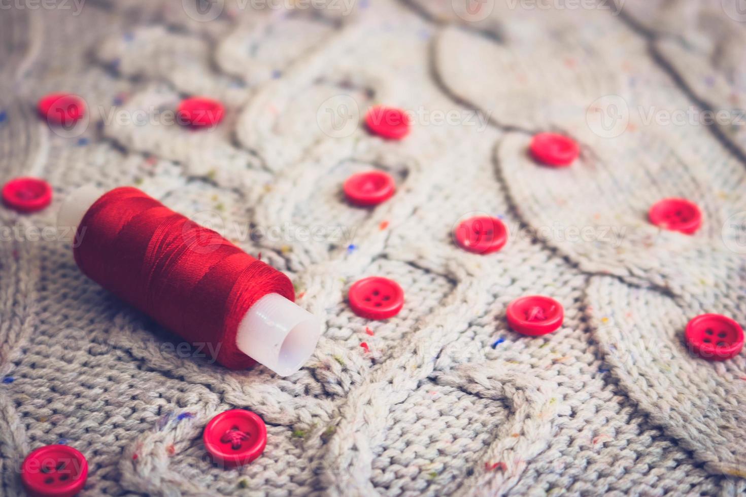 Beautiful texture of a soft warm natural sweater, fabrics with a knitted pattern of yarn and red small round buttons for sewing and a skein of red thread. The background photo