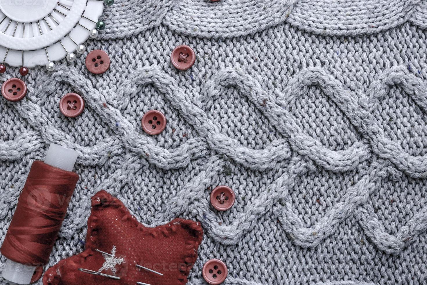 Beautiful texture of a soft warm natural sweater, fabrics with a knitted pattern and red small round buttons for sewing and a skein of thread, needle bed and needle pad. Flat lay. The background photo