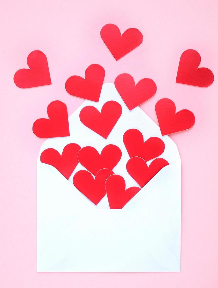 Red hearts and white envelope on pink background. Romantic message, letter, gift, vertical greeting card of Valentine's Day concept. Holiday festive creative design love composition. Flatlay, top view photo