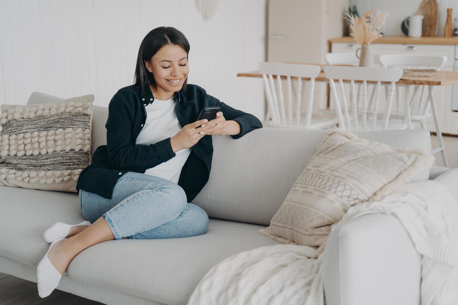 Smiling female uses phone apps, chatting in social networks, shopping online, sits on couch at home photo