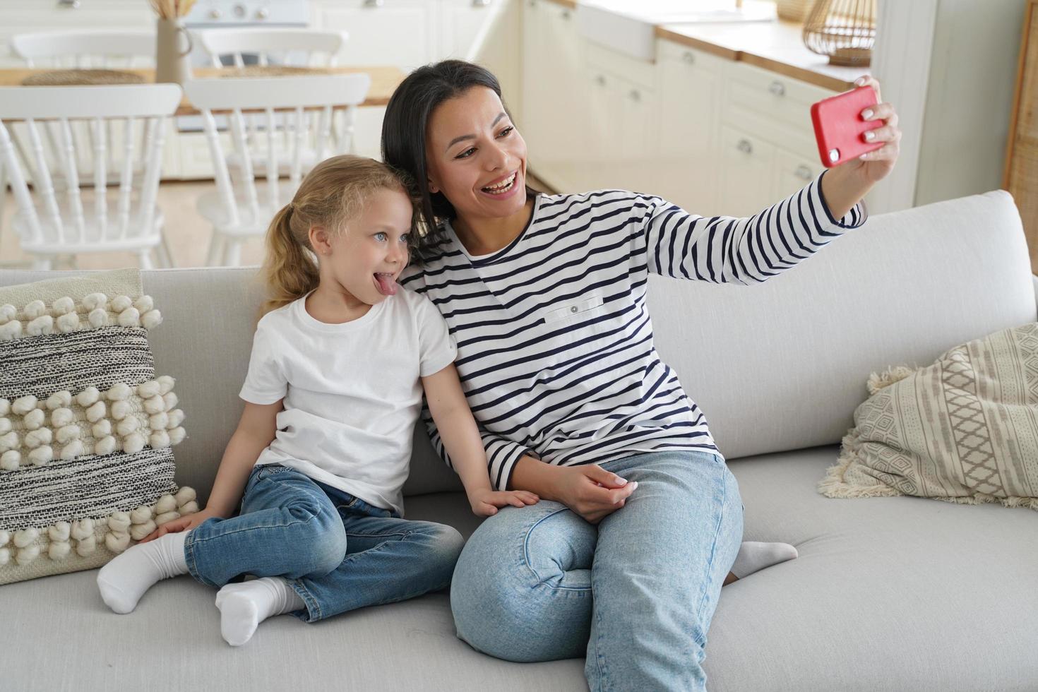 madre, hija pequeña adoptada, mostrando la lengua, tomando selfie familiar juntos, usando el teléfono en el sofá foto
