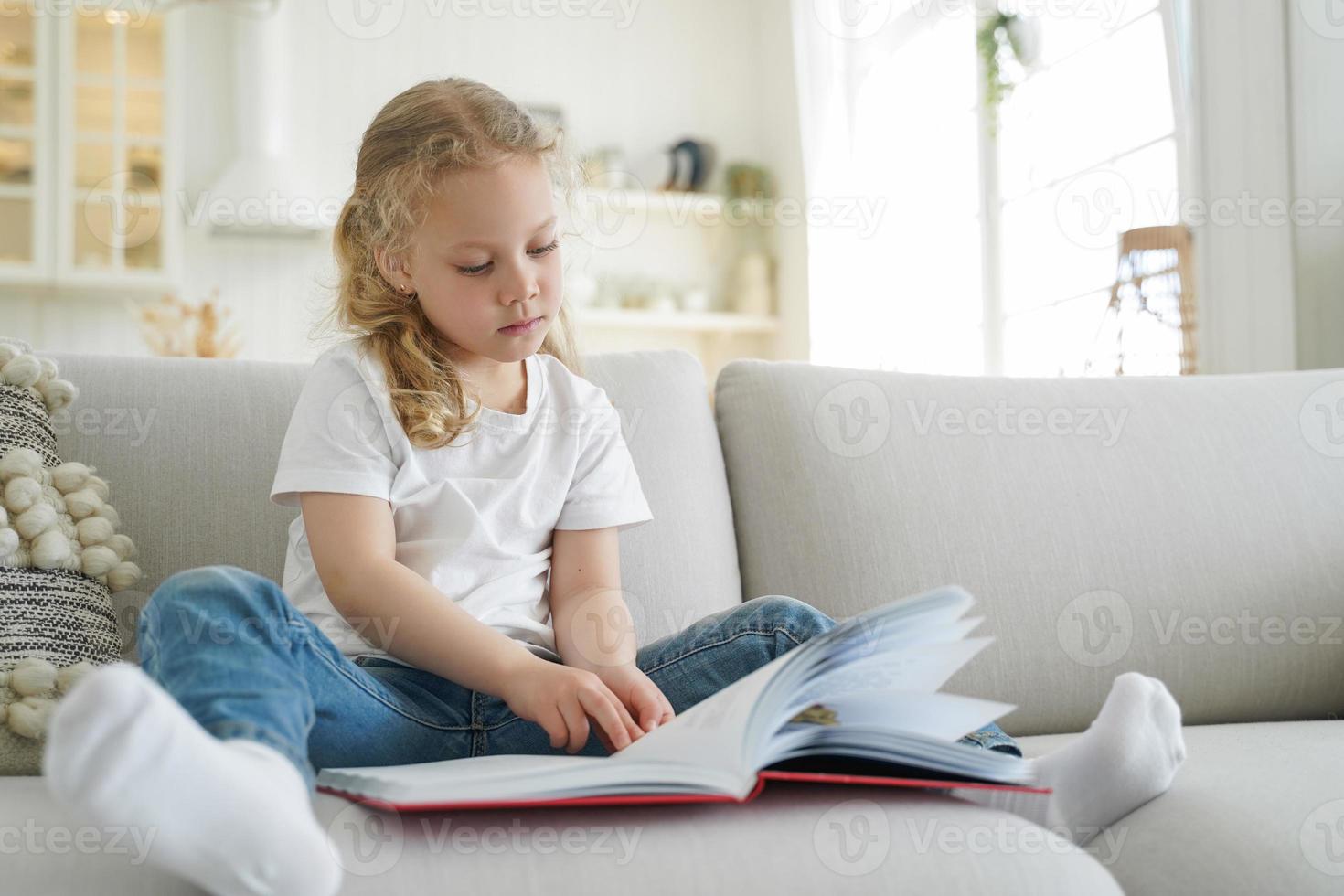 Little elementary child girl reads book, literature bestseller for children, sitting on sofa at home photo