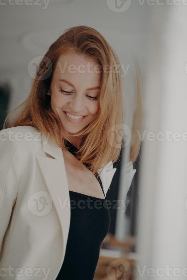 Smiling young woman with stylish haircut in white suit jacket posing near mirror. Female beauty photo