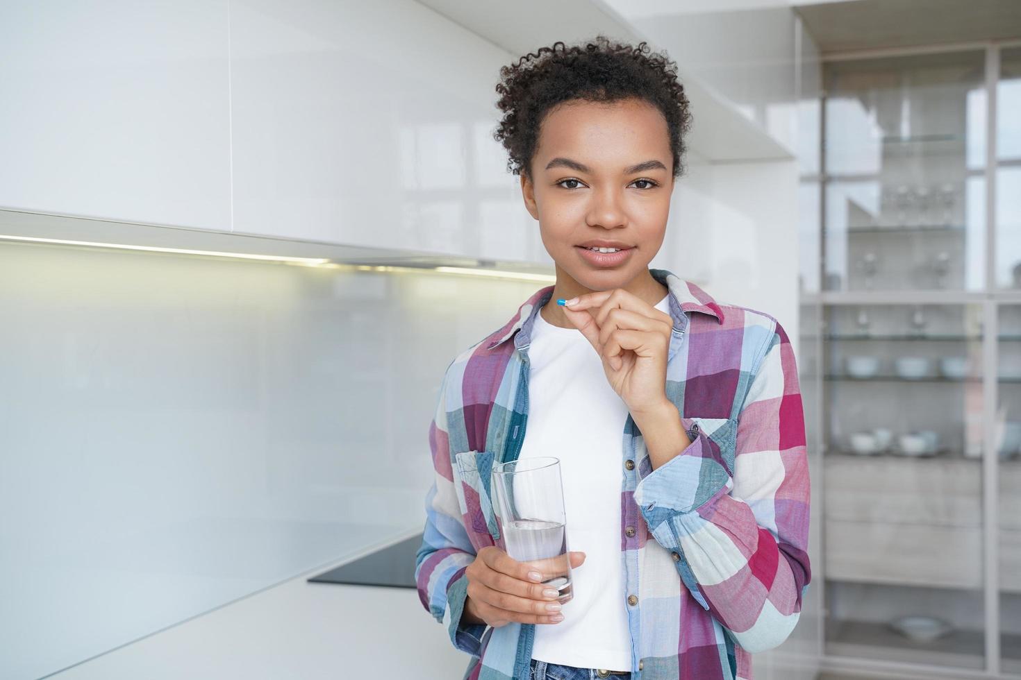 Mixed race young girl takes pills, vitamins, dietary supplement for wellness holding glass of water photo