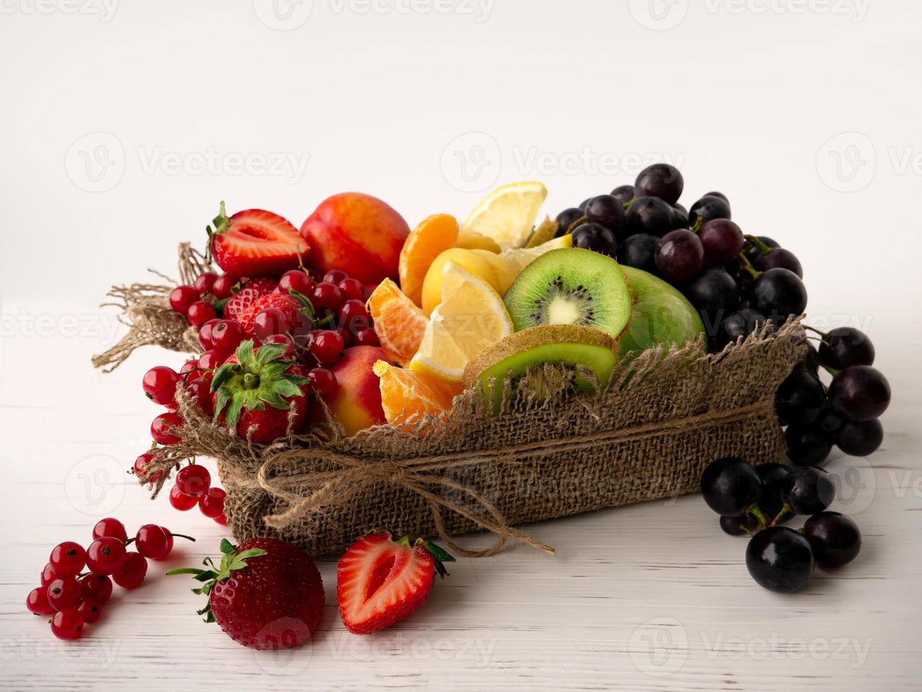 A mix of fresh fruits and berries of different colors in a burlap basket on a light background. Healthy summer food photo