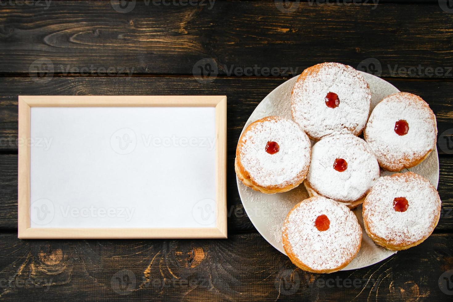 Happy Hanukkah. Empty frame for congratulations text and traditional dessert Sufganiyot on dark wooden background. Celebrating Jewish holiday. photo