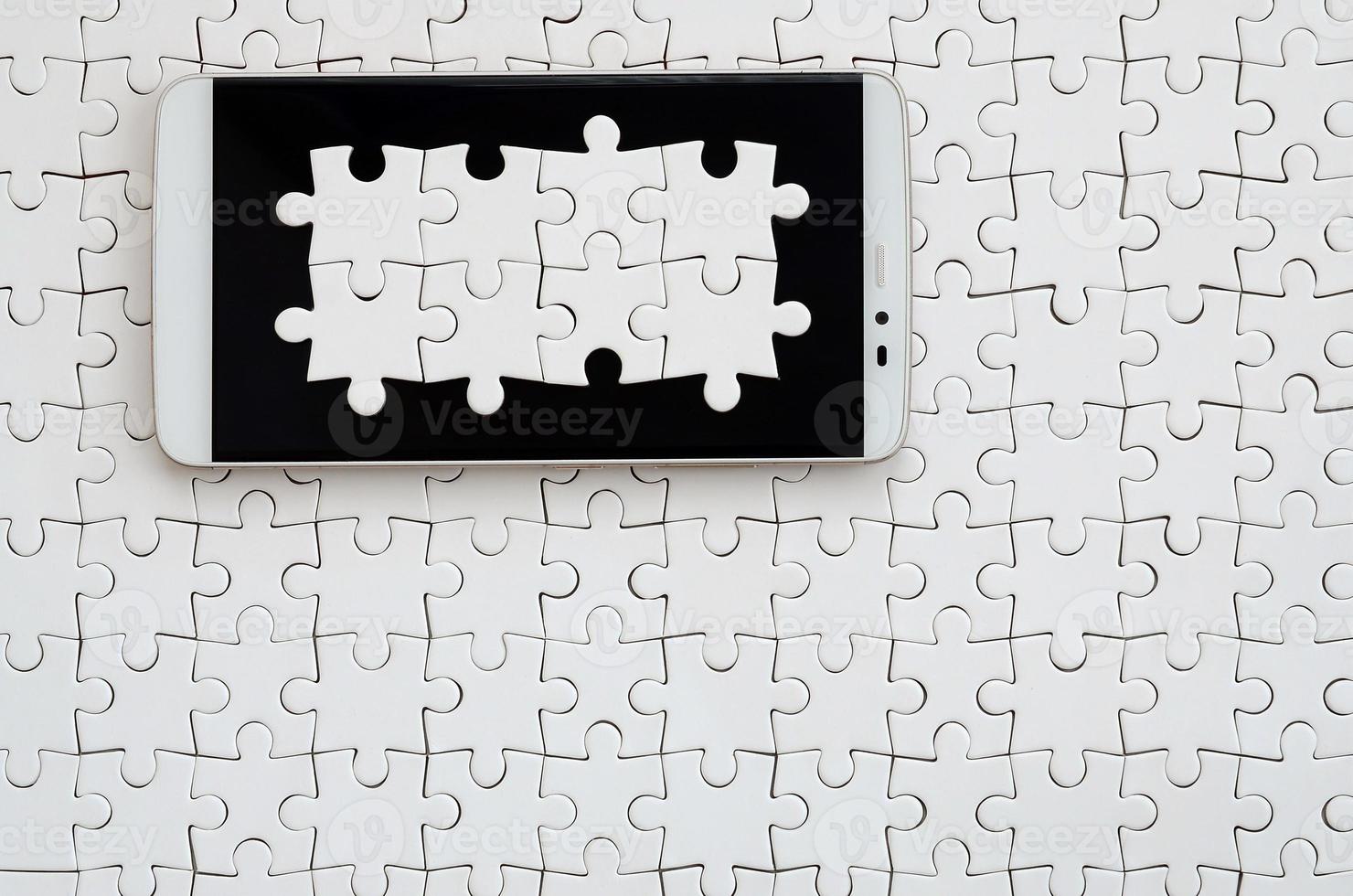 A modern big smartphone with several puzzle elements on the touch screen lies on a white jigsaw puzzle in an assembled state photo