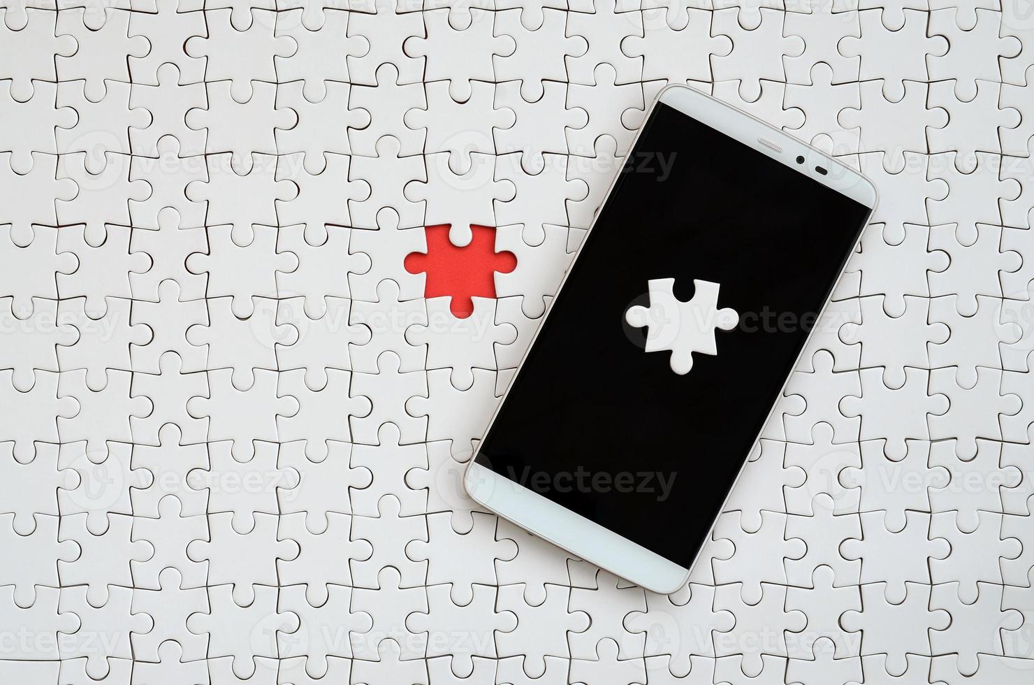 A modern big smartphone with several puzzle elements on the touch screen lies on a white jigsaw puzzle in an assembled state with missing elements photo