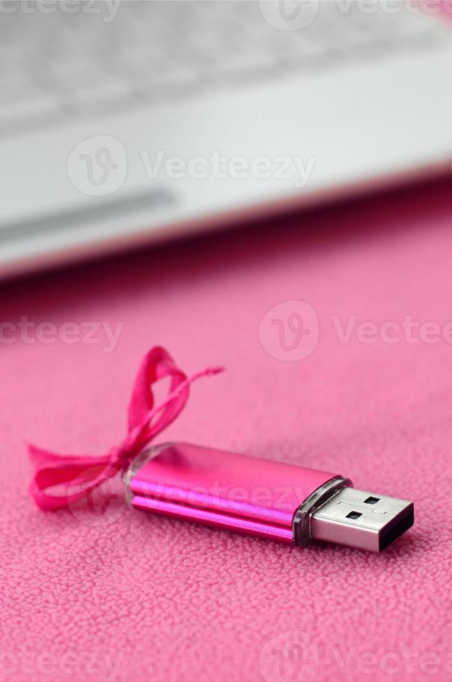 Brilliant pink usb flash memory card with a pink bow lies on a blanket of soft and furry light pink fleece fabric beside to a white laptop. Classic female gift design for a memory card photo