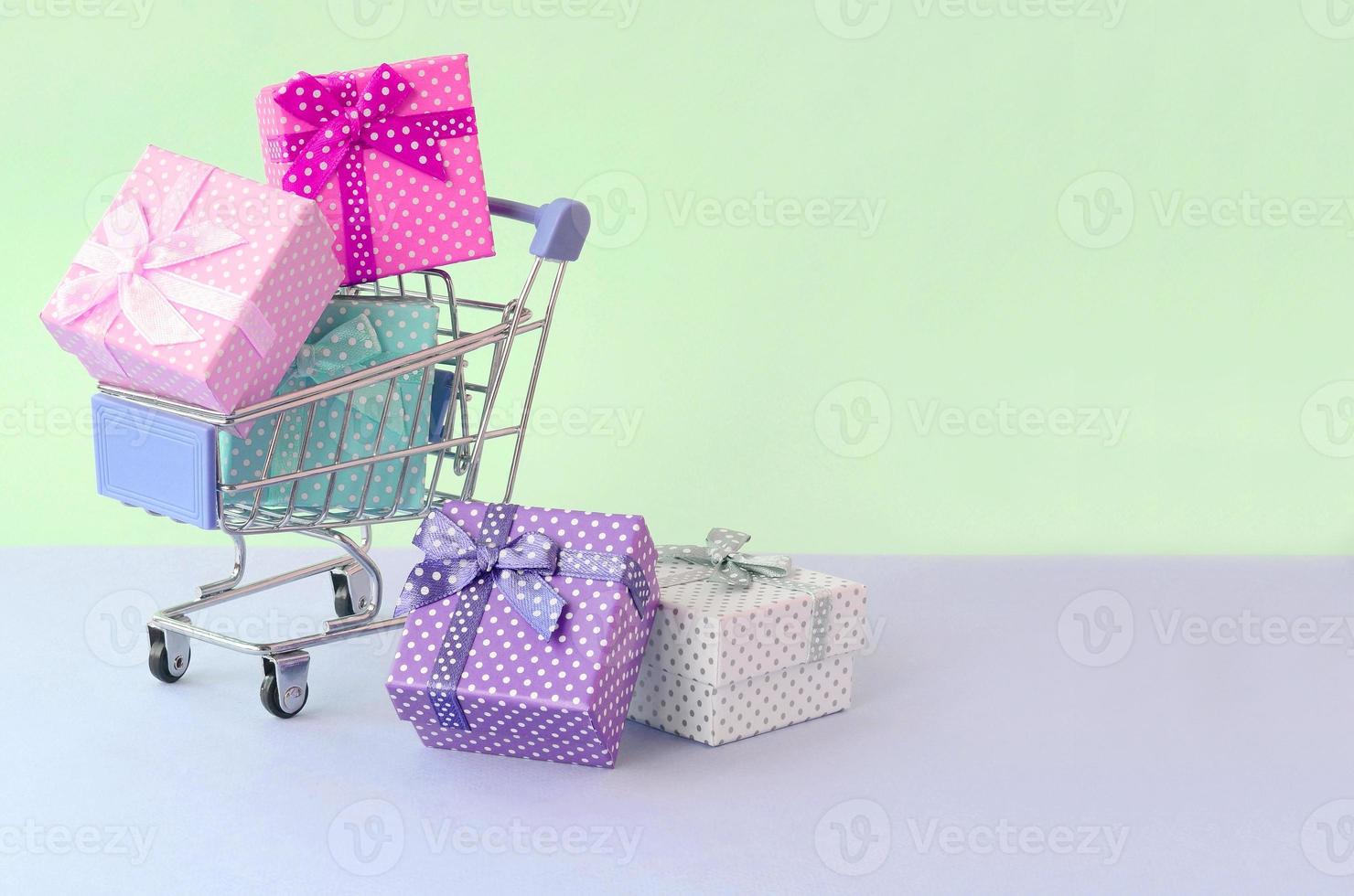 pequeñas cajas de regalo de diferentes colores con cintas en carrito de compras sobre un fondo violeta y azul pastel foto