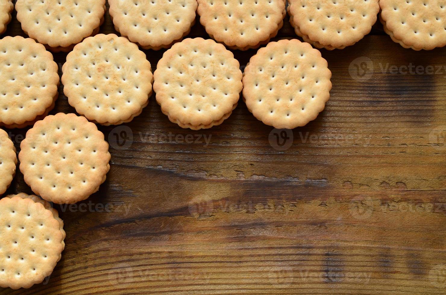 A round sandwich cookie with coconut filling lies in large quantities on a brown wooden surface. Photo of edible treats on a wooden background with copy space