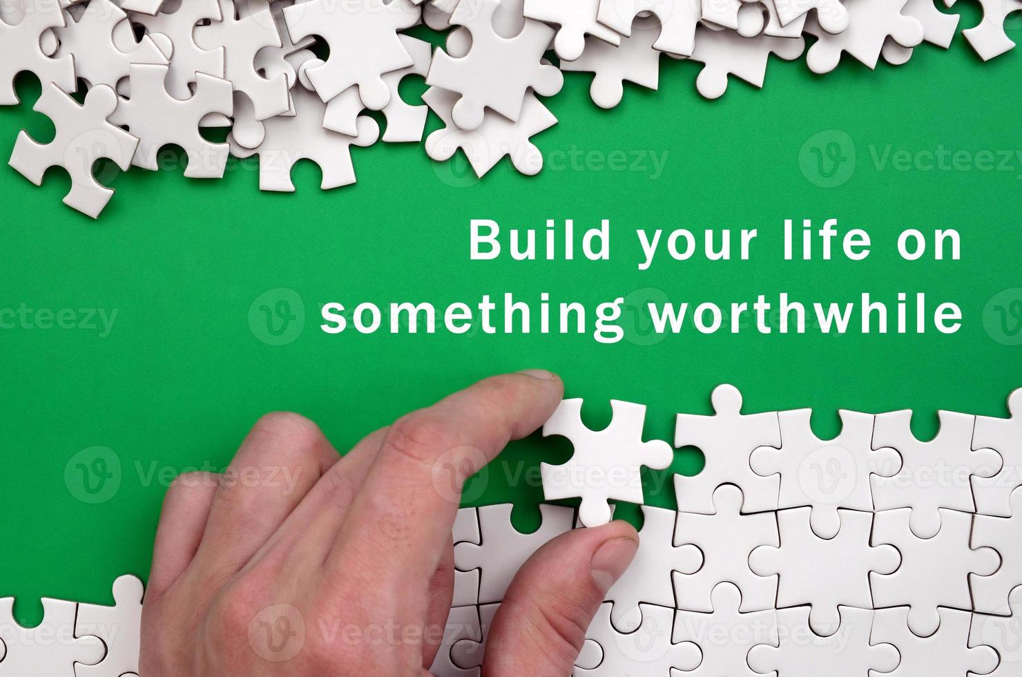 Build your life on something worthwhile. The hand folds a white jigsaw puzzle and a pile of uncombed puzzle pieces lies against the background of the green surface photo