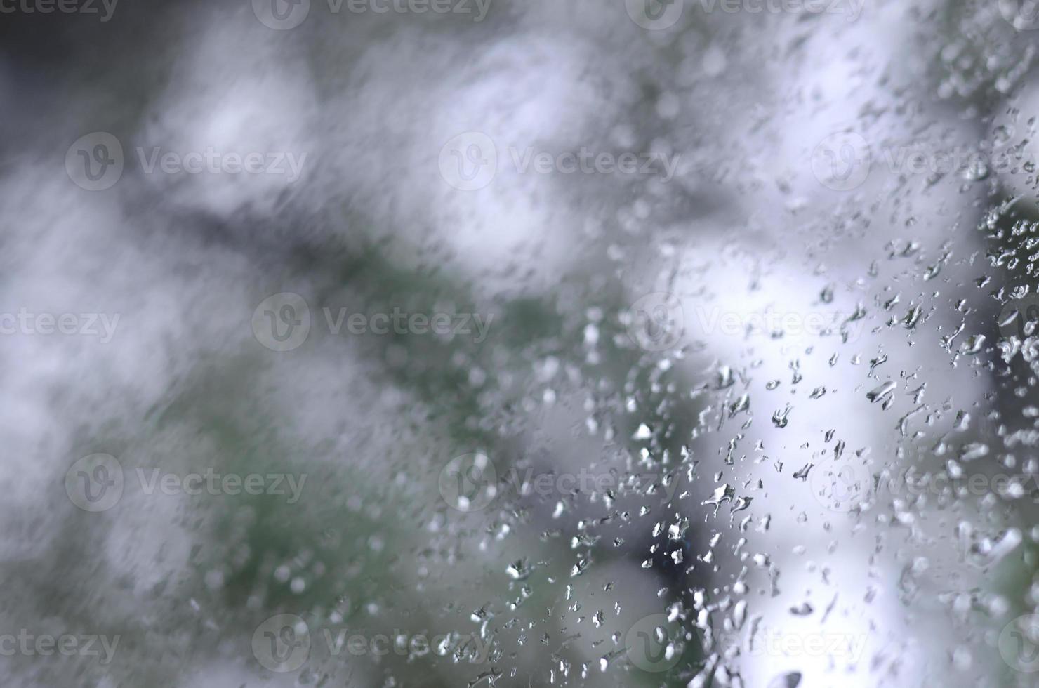 A photo of rain drops on the window glass with a blurred view of the blossoming green trees. Abstract image showing cloudy and rainy weather conditions