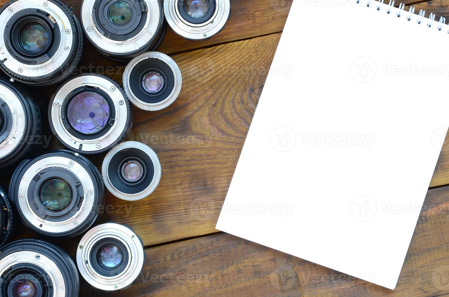 Several photographic lenses and white notebook lie on a brown wooden background. Space for text photo