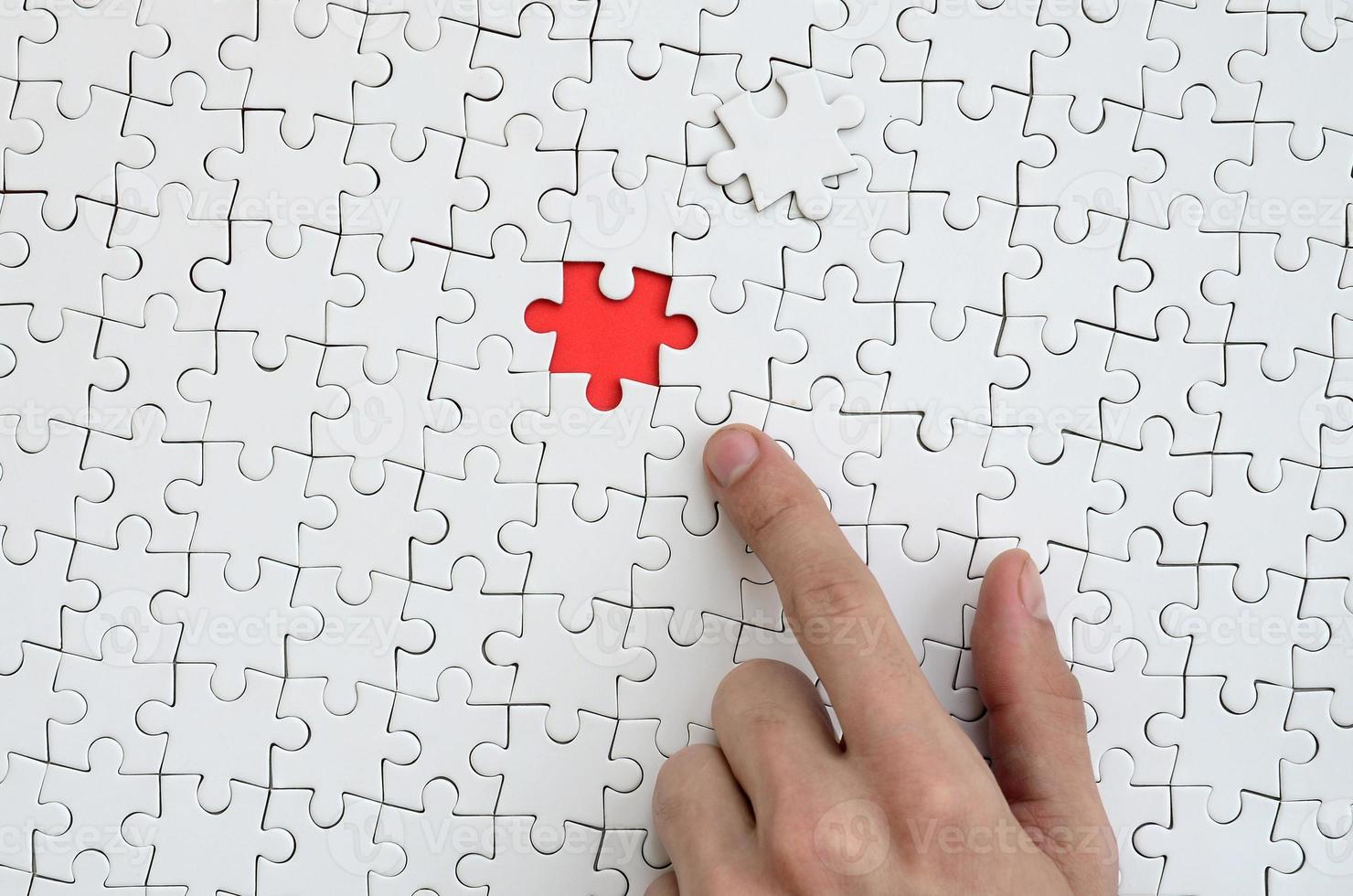 The texture of a white jigsaw puzzle in the assembled state with one missing element, forming a red space, pointed to by the finger of the male hand photo